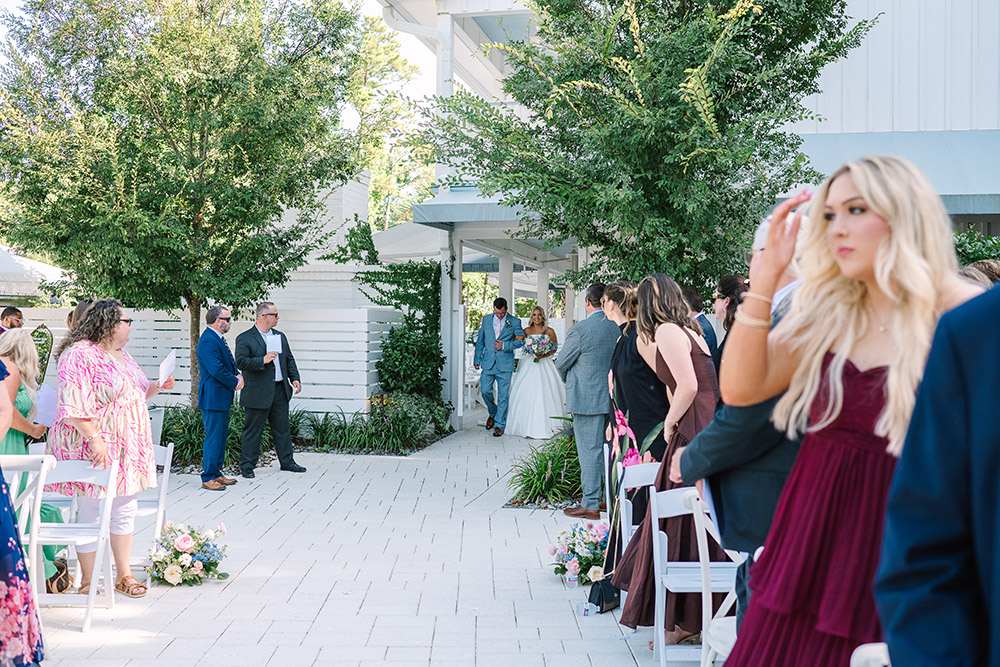 Bride walks down the aisle escorted by an older man during an outdoor wedding ceremony in front of a white chapel.