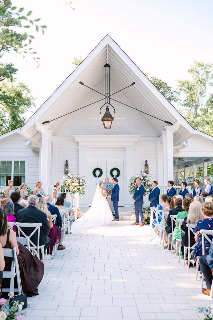 Bride and groom exchange vows at the altar during an outdoor chapel ceremony surrounded by guests and pastel floral arrangements.