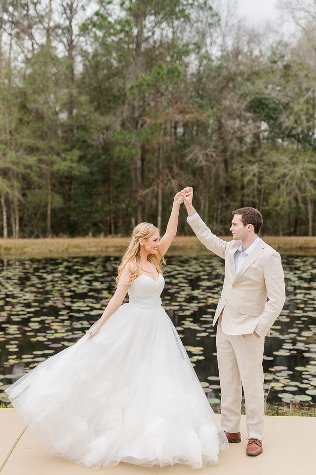 Bride twirling in strapless tulle wedding gown beside pond with lily pads, groom in beige suit