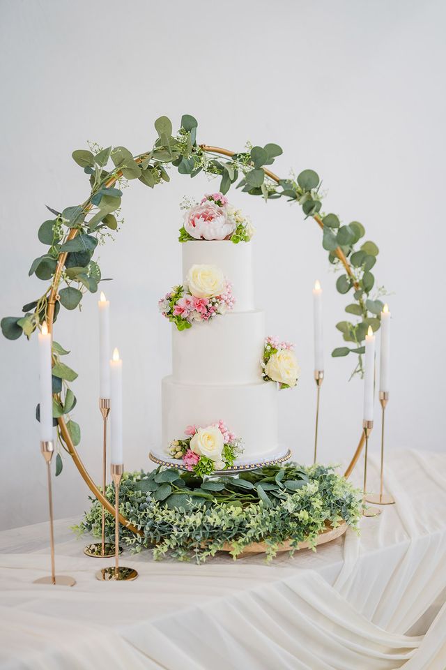 Three-tier white wedding cake with blush and ivory florals displayed beneath circular greenery arch with candles