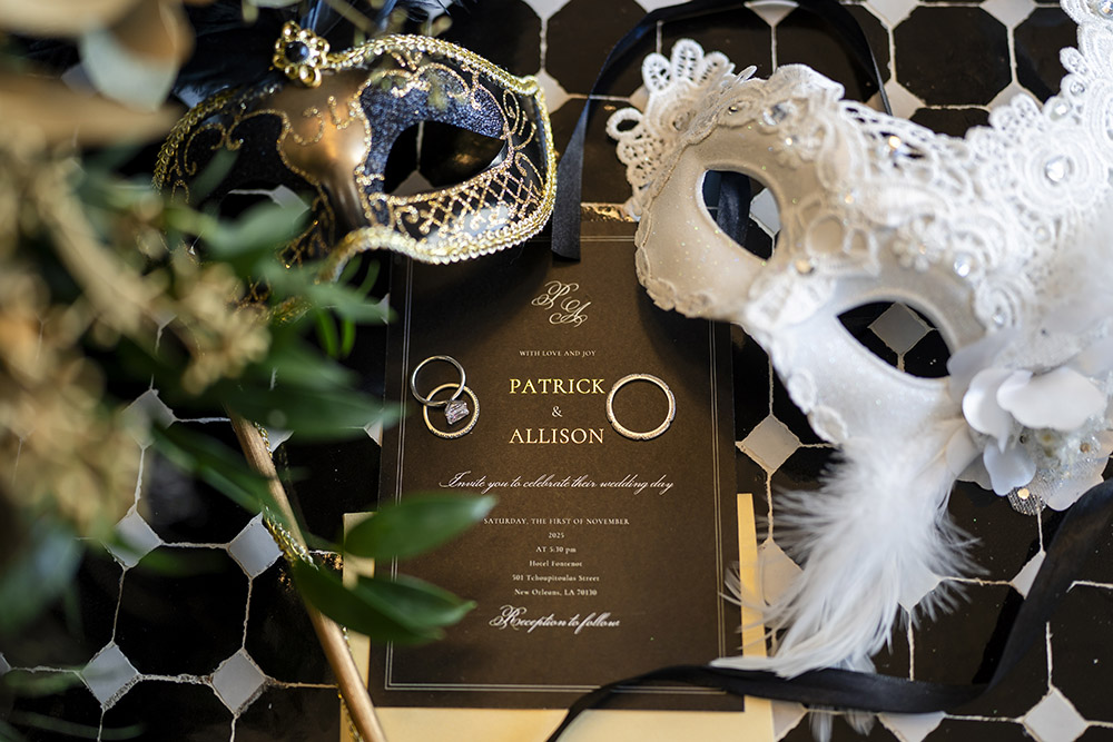 Black and gold wedding invitation suite styled with masquerade masks, wedding bands, and elegant ribbon details on a patterned tile floor.