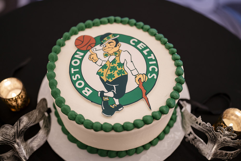 Groom’s cake decorated with the Boston Celtics logo displayed alongside ornate silver masquerade masks and candlelight accents.
