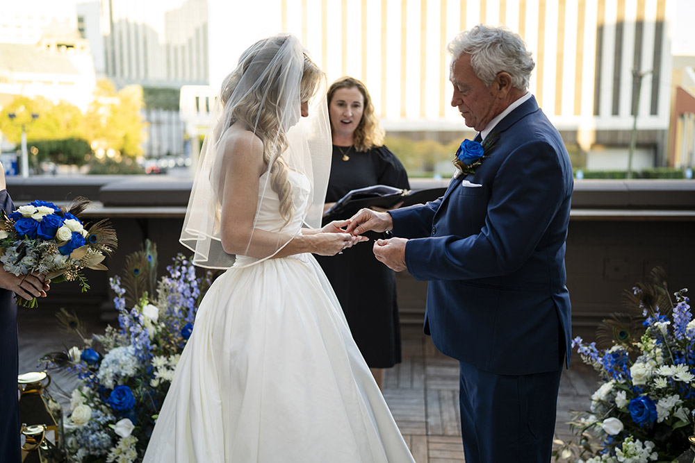 Bride and groom exchange rings during an intimate rooftop wedding ceremony surrounded by lush blue and white floral arrangements.