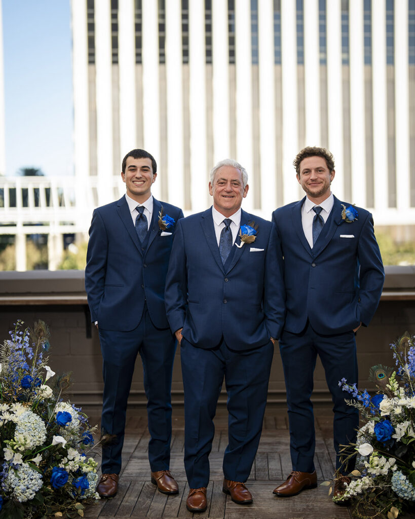 Groom poses with two groomsmen in coordinated navy blue suits during rooftop wedding portraits framed by blue and white ceremony florals.
