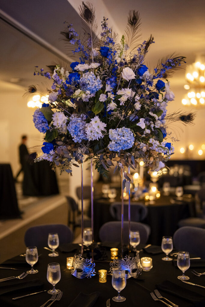 Tall reception centerpiece overflowing with blue roses, hydrangeas, white blooms, and peacock feathers rises above candlelit reception tables.
