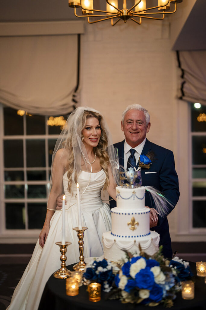 Bride and groom smile beside their elegant white wedding cake decorated with blue beading, gold fleur-de-lis accents, and masquerade-inspired details.