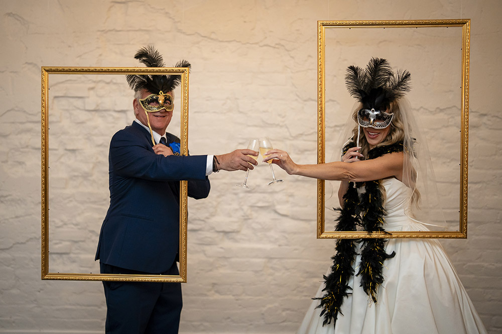 Bride and groom wearing ornate masquerade masks toast with champagne while posing inside suspended gold picture frames at their reception photo booth.