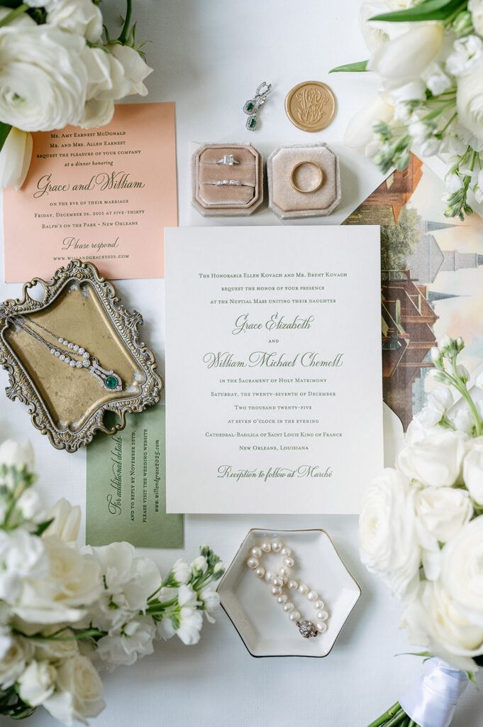 Flat lay of Grace and William’s wedding invitations styled with emerald jewelry, velvet ring boxes, pearl bracelet, wax seal, and white floral arrangements on a soft ivory backdrop.