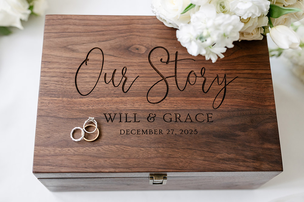 Custom wooden wedding keepsake box engraved with “Our Story,” Will & Grace’s names, and wedding date, accented with wedding rings and white flowers.
