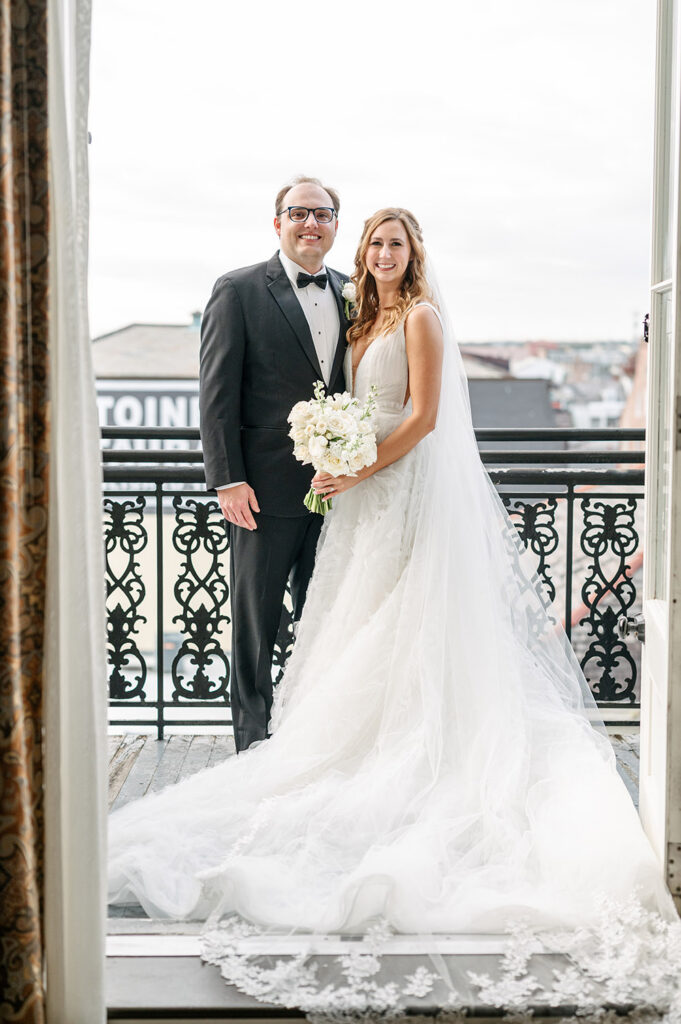Bride and groom posing on a French Quarter balcony in New Orleans, the bride wearing a flowing layered tulle gown and cathedral veil beside the groom in a classic black tuxedo.