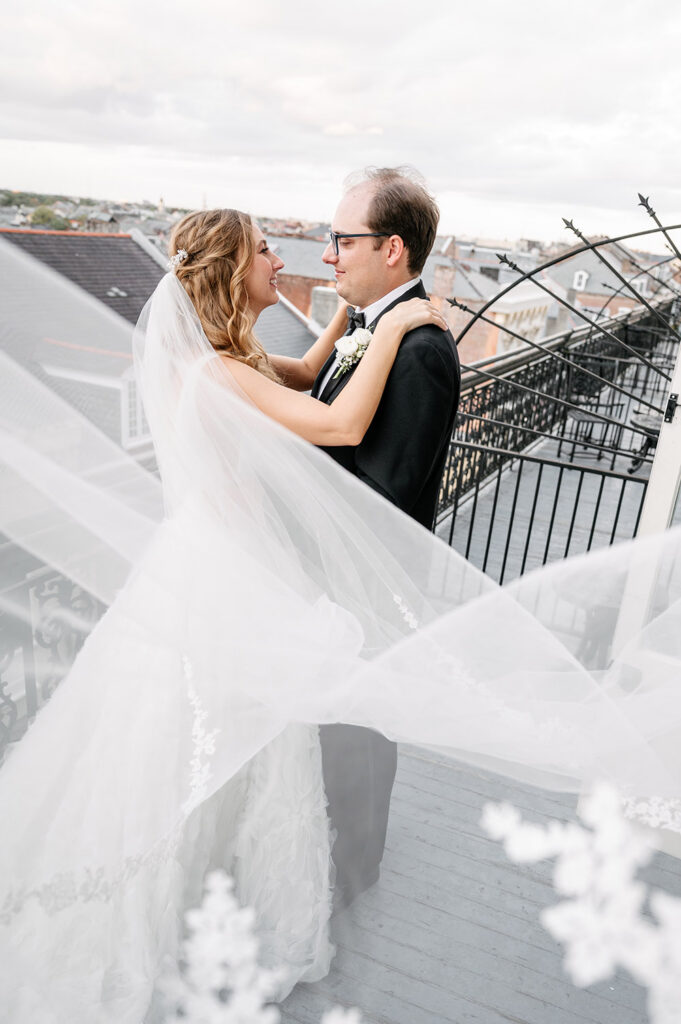 Romantic balcony portrait of the newlyweds embracing beneath the bride’s windswept cathedral veil overlooking the New Orleans skyline.