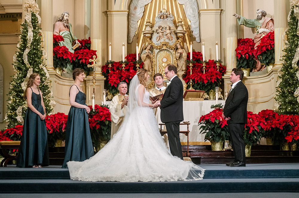 Bride and groom exchanging vows at the altar during a Christmas-season ceremony surrounded by poinsettias, candlelight, and attendants in deep green attire.