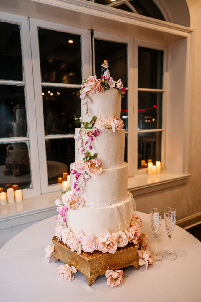 Four-tier white wedding cake decorated with cascading blush sugar flowers and displayed beside champagne flutes and candlelight at the reception.