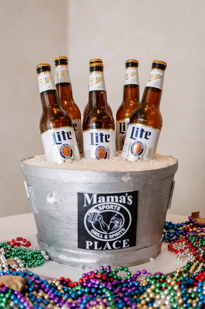 Playful groom's cake made to look like Miller Lite bottles chilling in an ice bucket labeled “Mama’s Place” surrounded by colorful Mardi Gras beads
