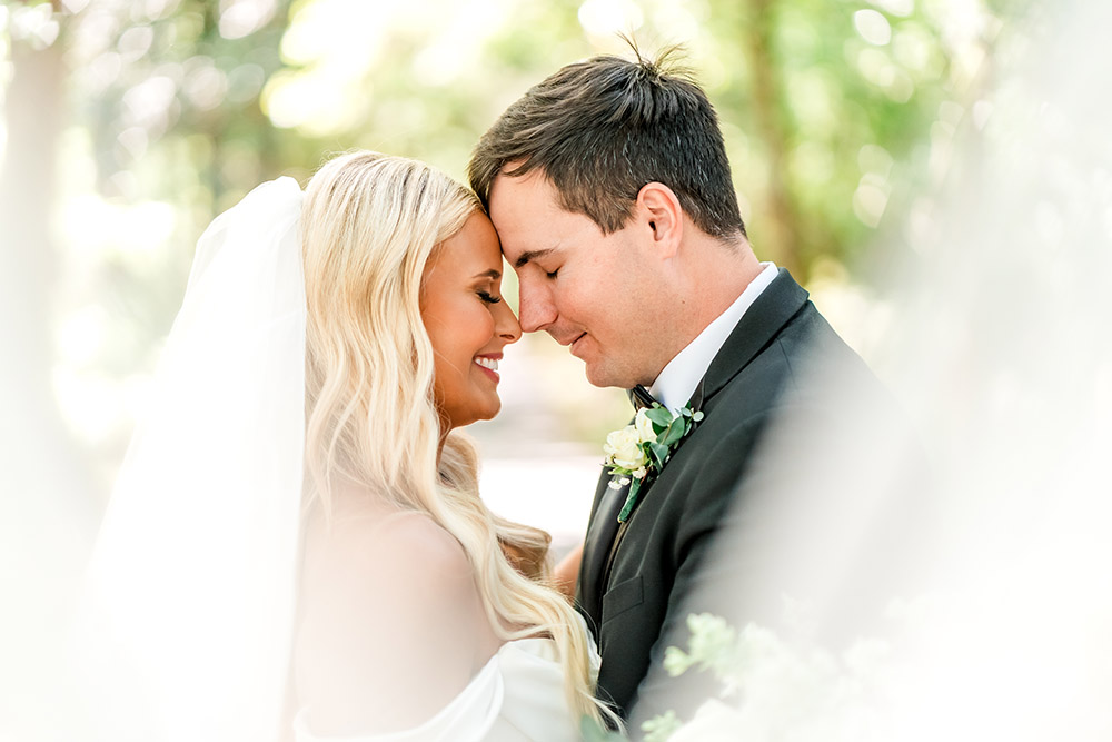 Bride and groom smile with their foreheads touching during an intimate portrait framed by soft greenery and the bride’s flowing veil.