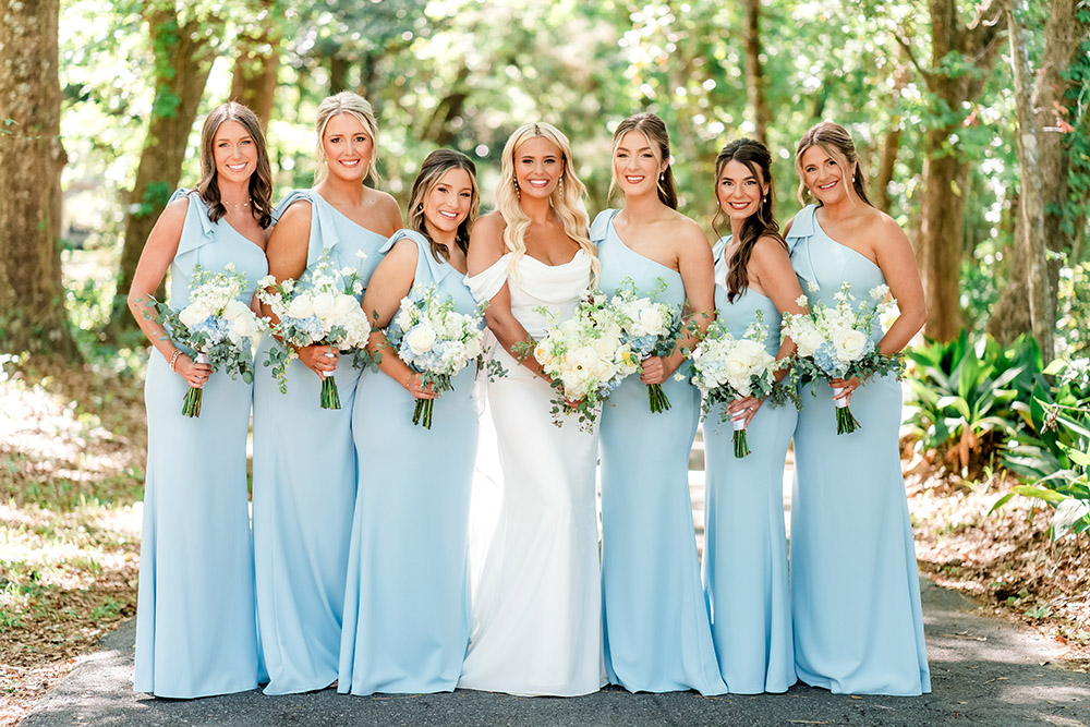Bride poses with six bridesmaids in matching light blue one-shoulder gowns, each holding white and blue floral bouquets beneath towering oak trees.