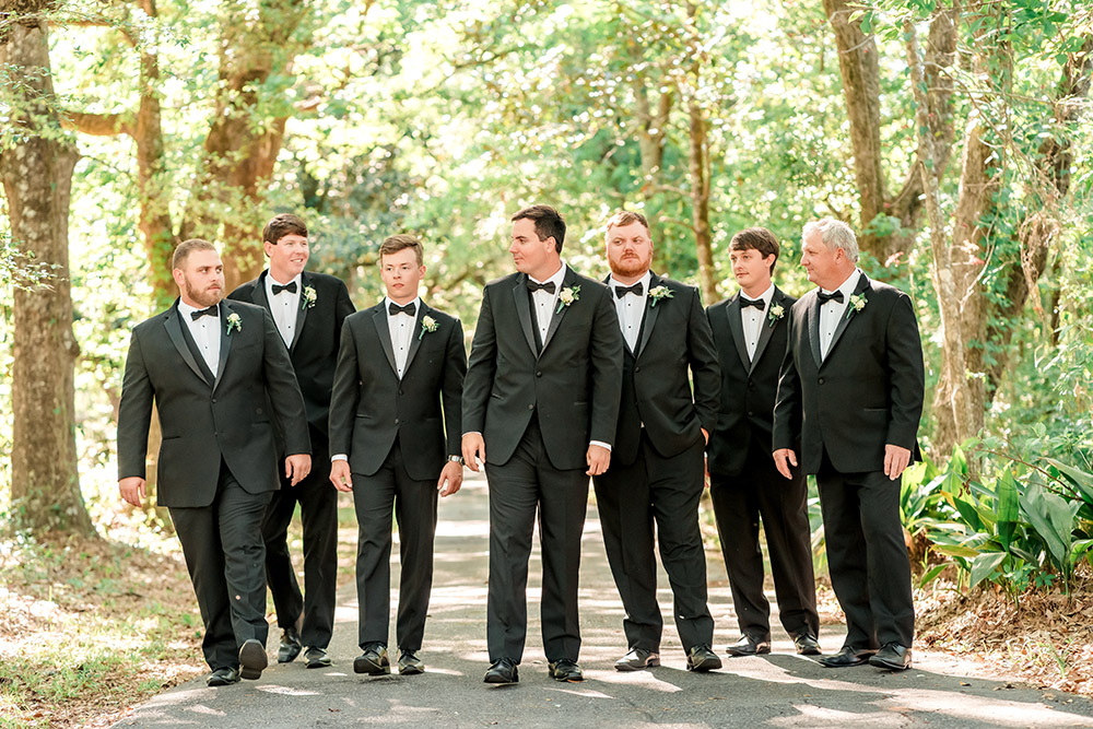 Groom walks with his groomsmen along a shaded tree-lined path, all dressed in classic black tuxedos with white rose boutonnieres.