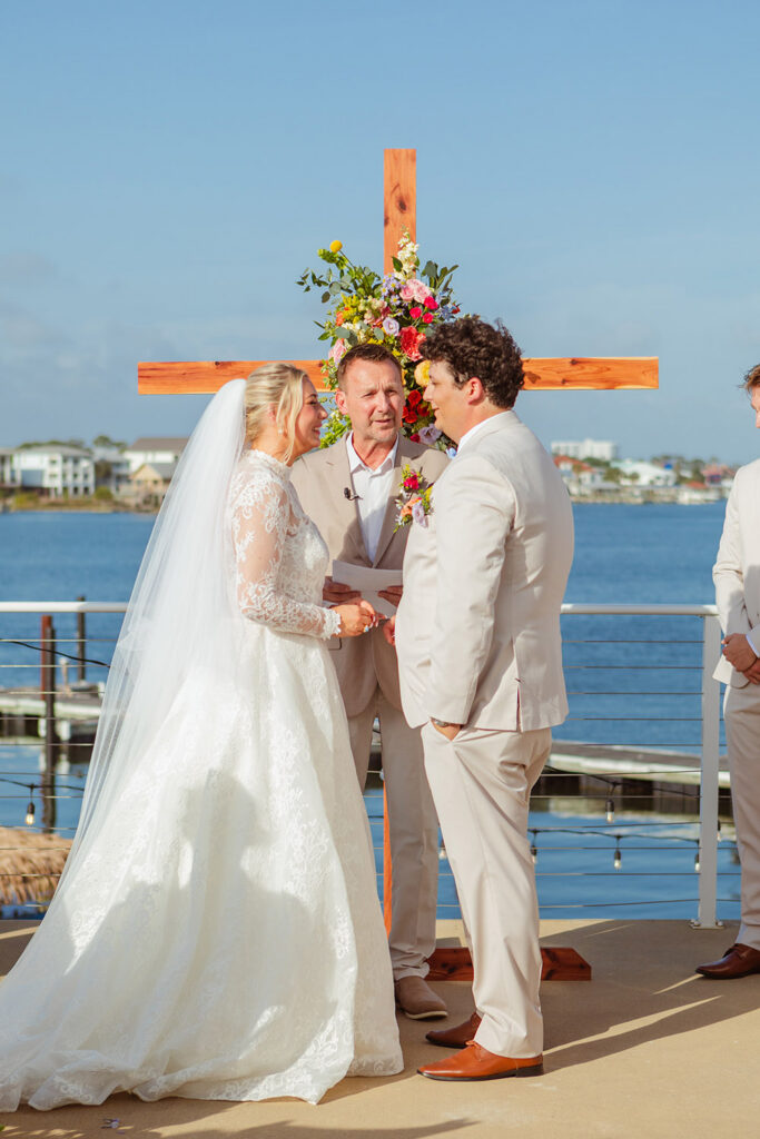 The couple exchanges vows during an outdoor waterfront ceremony beneath a wooden cross decorated with colorful blooms.