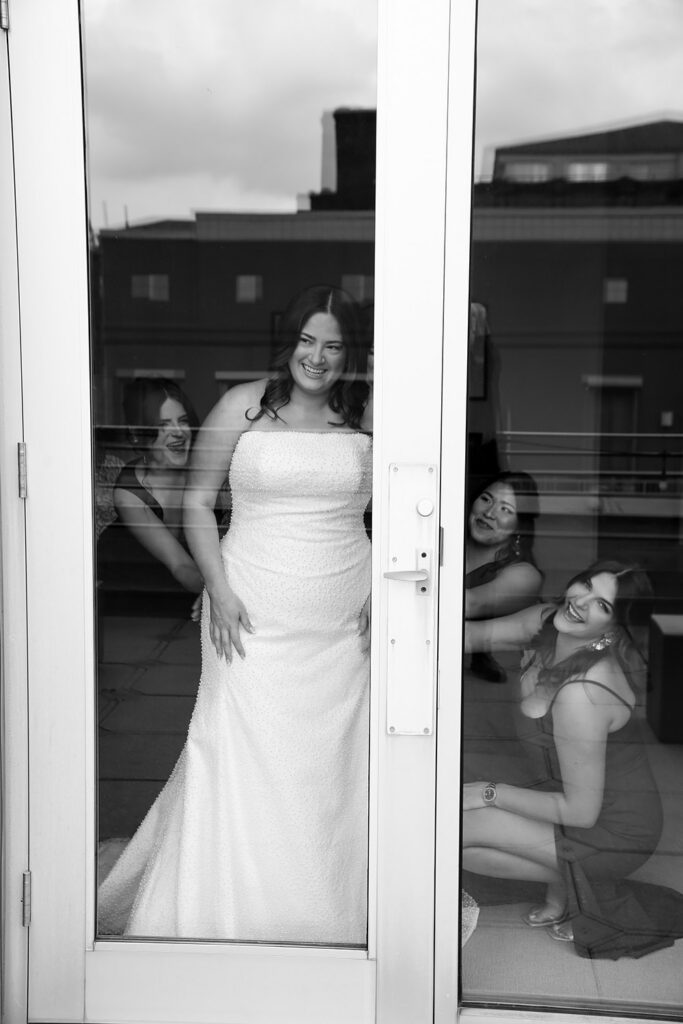 Bride smiles while her bridesmaids peek around a glass door before the ceremony.