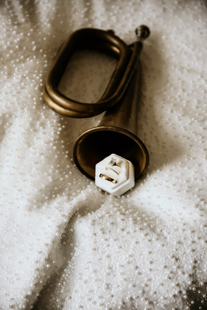 Bride's engagement ring and wedding band displayed in a white velvet ring box beside a vintage brass horn on beaded fabric.