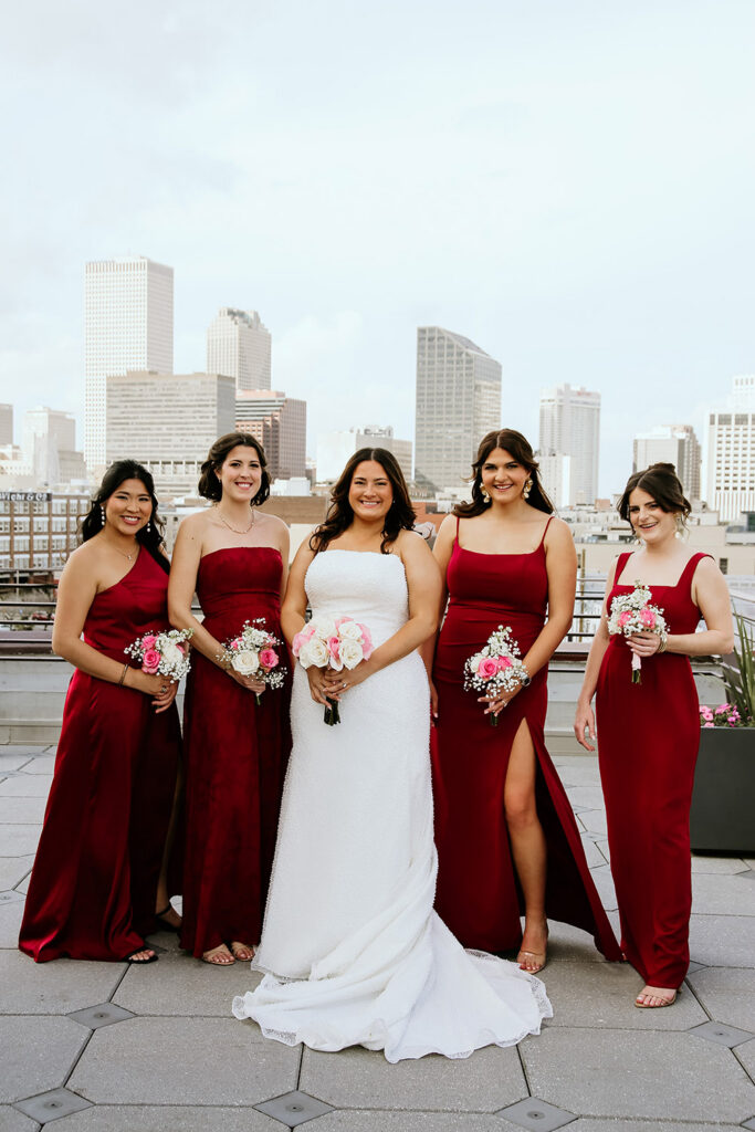 Bride poses with five bridesmaids in burgundy dresses on a rooftop overlooking downtown New Orleans.