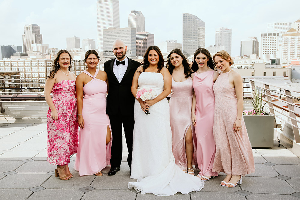 Bride and groom pose on a rooftop with five women in pink dresses, with the New Orleans skyline in the background.