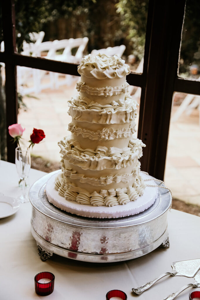 Four-tier white wedding cake decorated with dramatic ruffled buttercream displayed by a window.