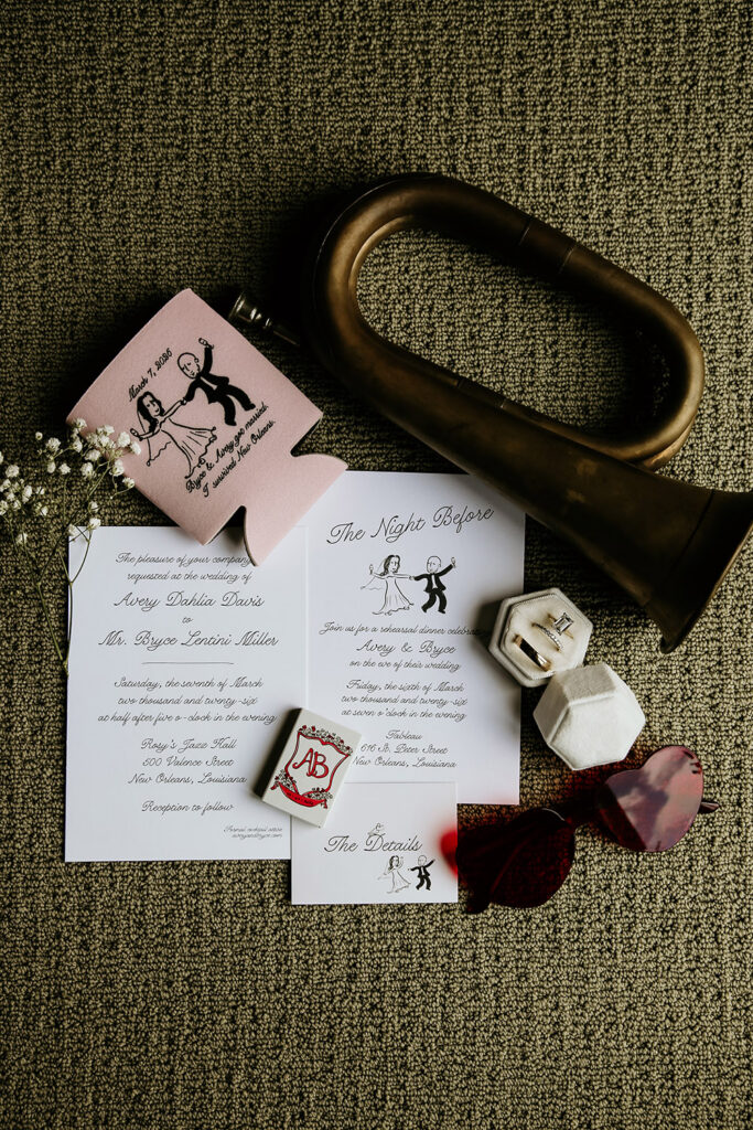 Wedding invitation suite with custom stationery, a vintage horn, ring box, sunglasses, and floral accents arranged in a flat lay.