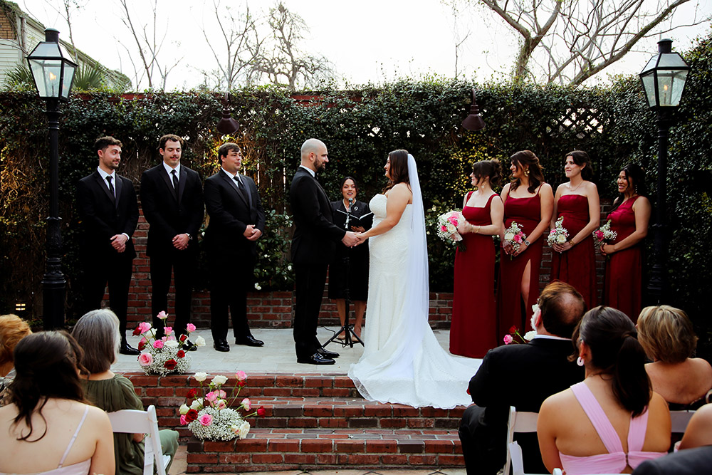 Bride and groom exchange vows during an outdoor courtyard ceremony with their wedding party standing beside them.