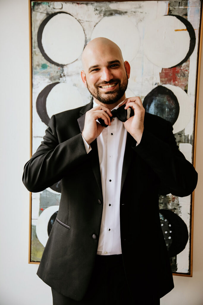 Groom adjusts his black bow tie while smiling in a classic black tuxedo.