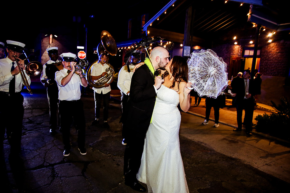 Bride and groom kiss during a nighttime second line parade led by a brass band.