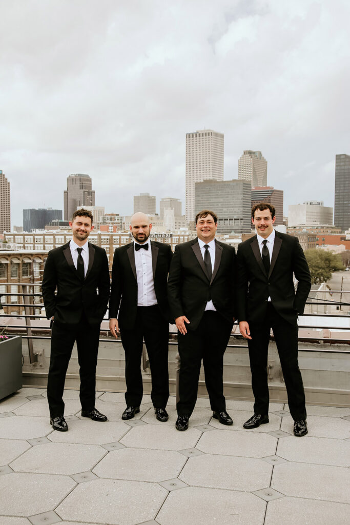 Groom and three groomsmen pose on a rooftop with the New Orleans skyline in the background.