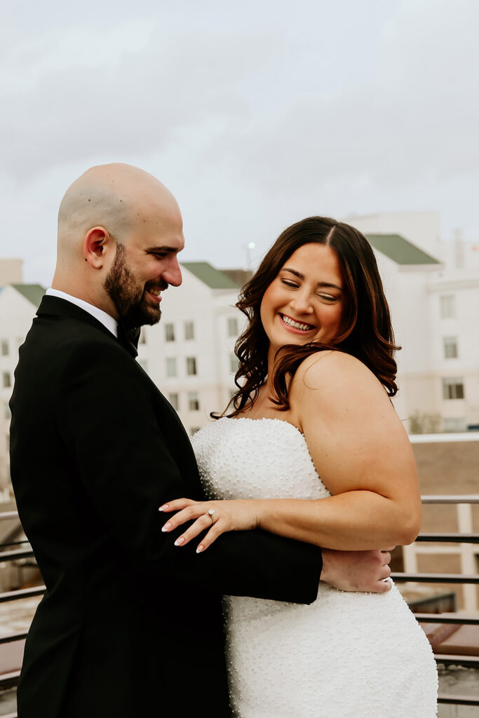 Bride smiles over her shoulder while embracing the groom on a rooftop before their ceremony.