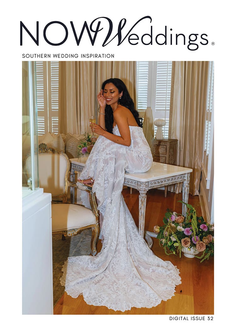 Bride in a lacy white gown smiles on a vanity, holding a glass, with flowers nearby. Text: NOW Weddings: Southern Wedding Inspiration.