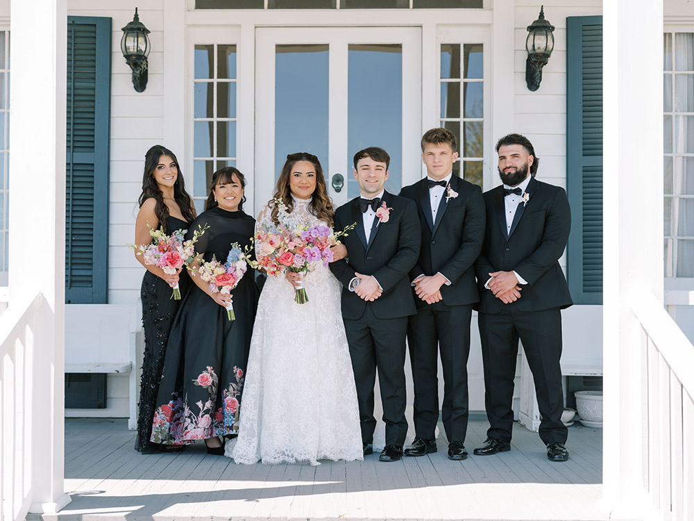 Bride and groom pose with their wedding party on the front porch, with the bride holding a vibrant bouquet of pink and peach blooms.