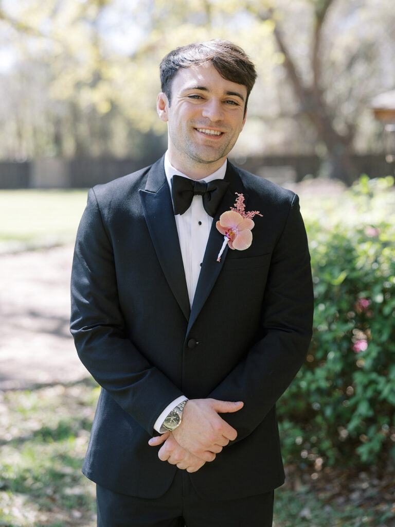 Groom in a classic black tuxedo with a blush orchid boutonniere smiling outdoors before the ceremony.