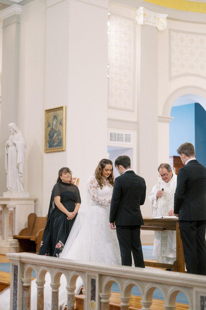 Bride and groom exchange rings during a Catholic wedding ceremony as the officiant looks on inside the church.