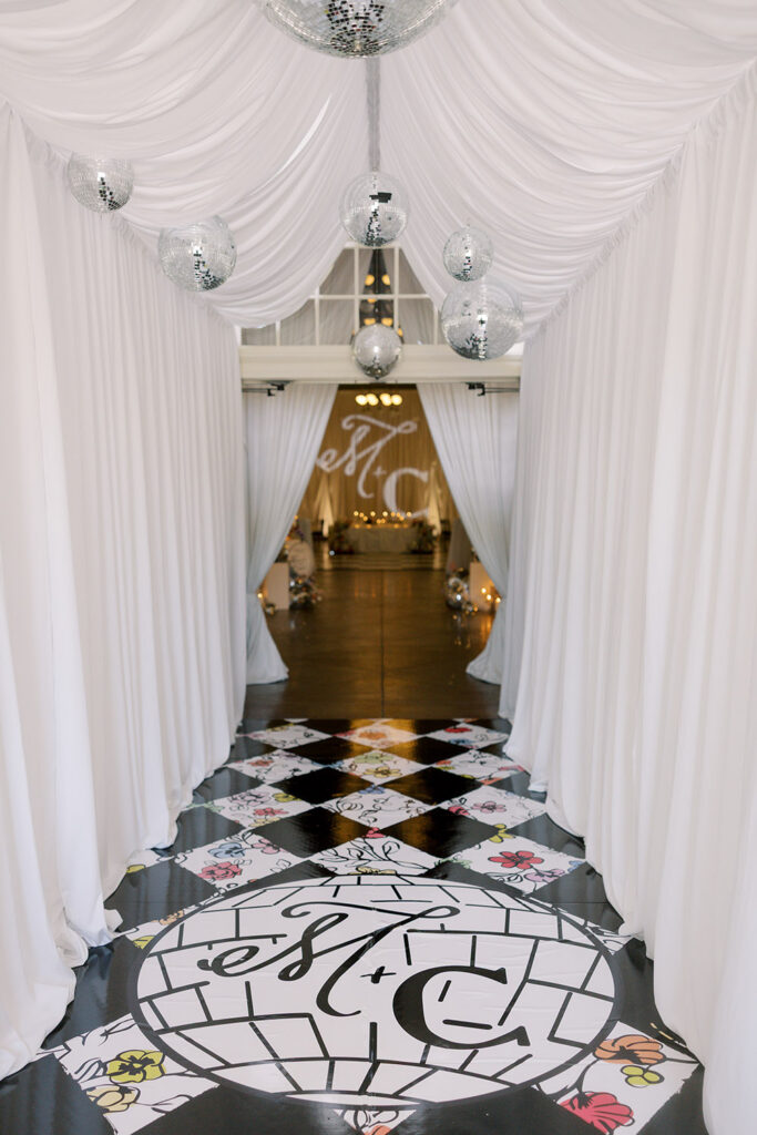 A draped white reception entrance lined with hanging disco balls leads into the ballroom, featuring the couple's custom monogram on the floor and backdrop.