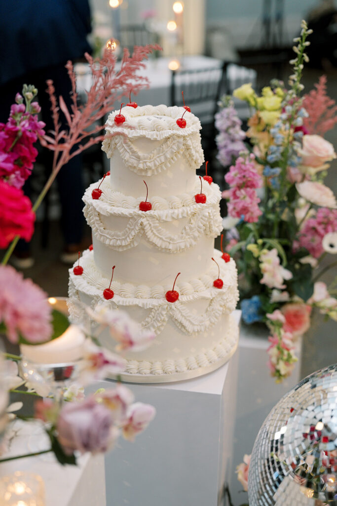 Three-tier white wedding cake with vintage-style ruffled buttercream and bright red cherries displayed among colorful floral arrangements.