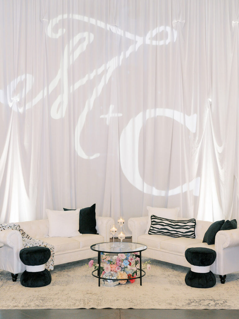 White lounge seating with black accent pillows sits beneath a large projection of the couple's monogram on a draped backdrop.