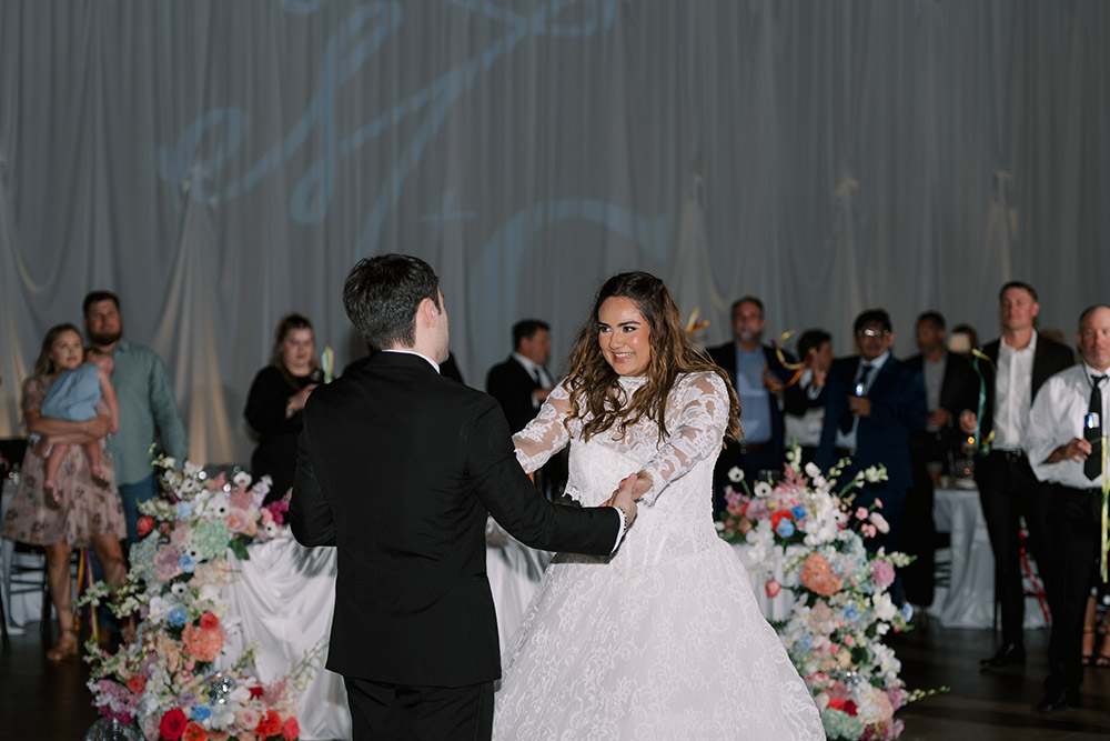 Bride and groom smile at one another during their first dance surrounded by guests and colorful floral arrangements
