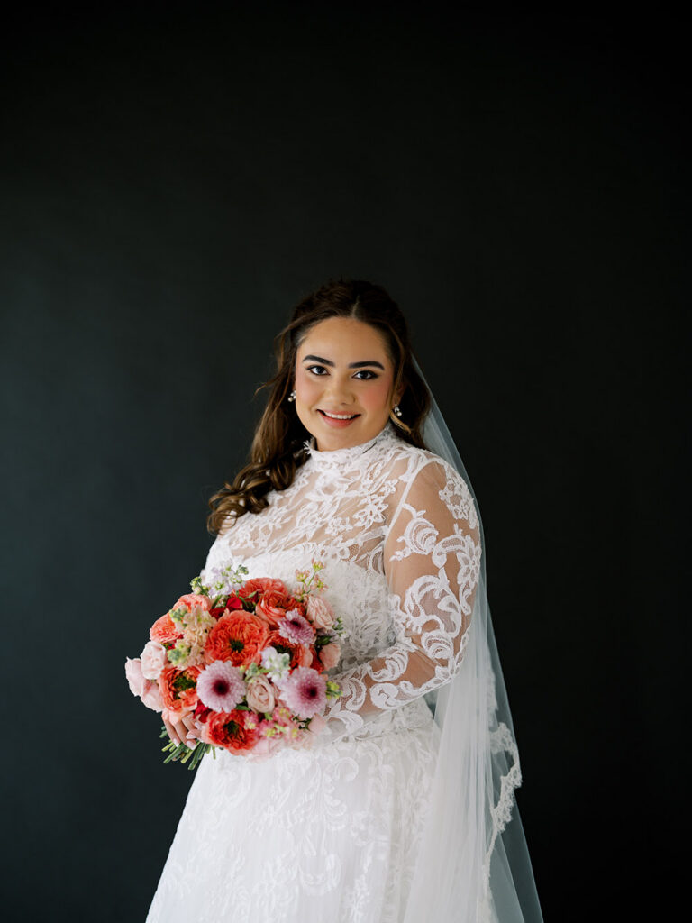 Bride in a high-neck lace wedding gown holds a bouquet of coral, blush, and pink flowers during a studio bridal portrait against a black backdrop.