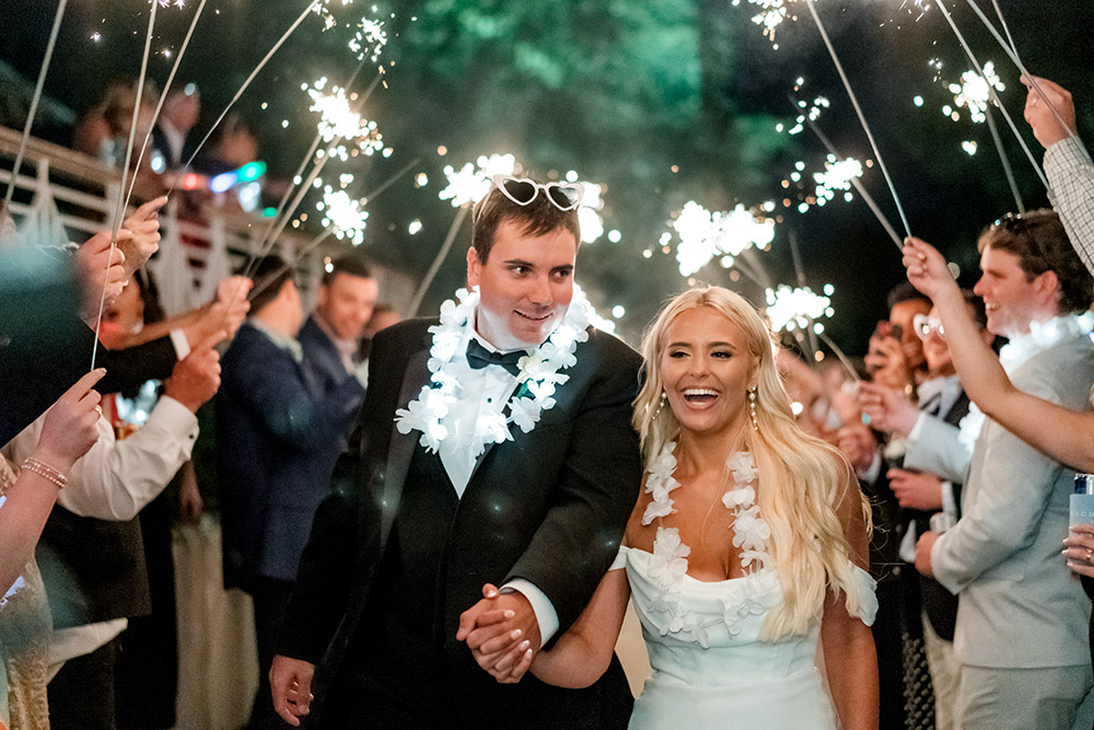 Newlyweds smile as they walk hand in hand through a sparkling send-off, wearing white floral leis and surrounded by cheering guests.