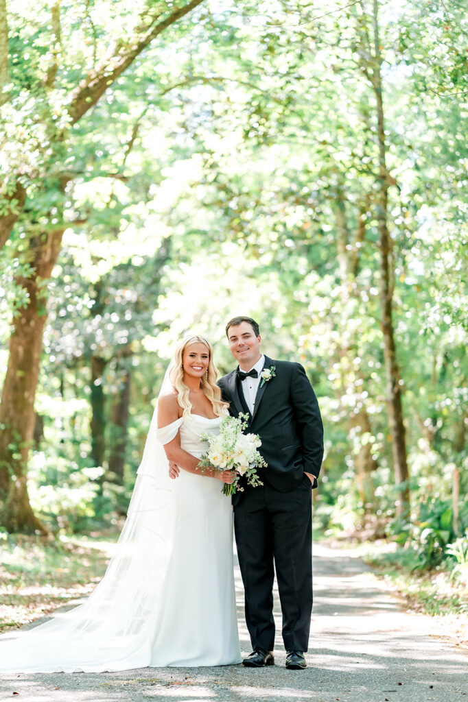 Bride and groom pose together on a tree-lined path, smiling while holding a lush white and blue wedding bouquet.