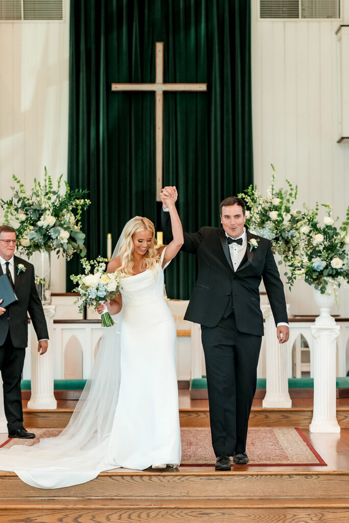 Newlyweds celebrate their marriage by walking hand in hand down the church aisle beneath a large wooden cross and elegant floral arrangements.