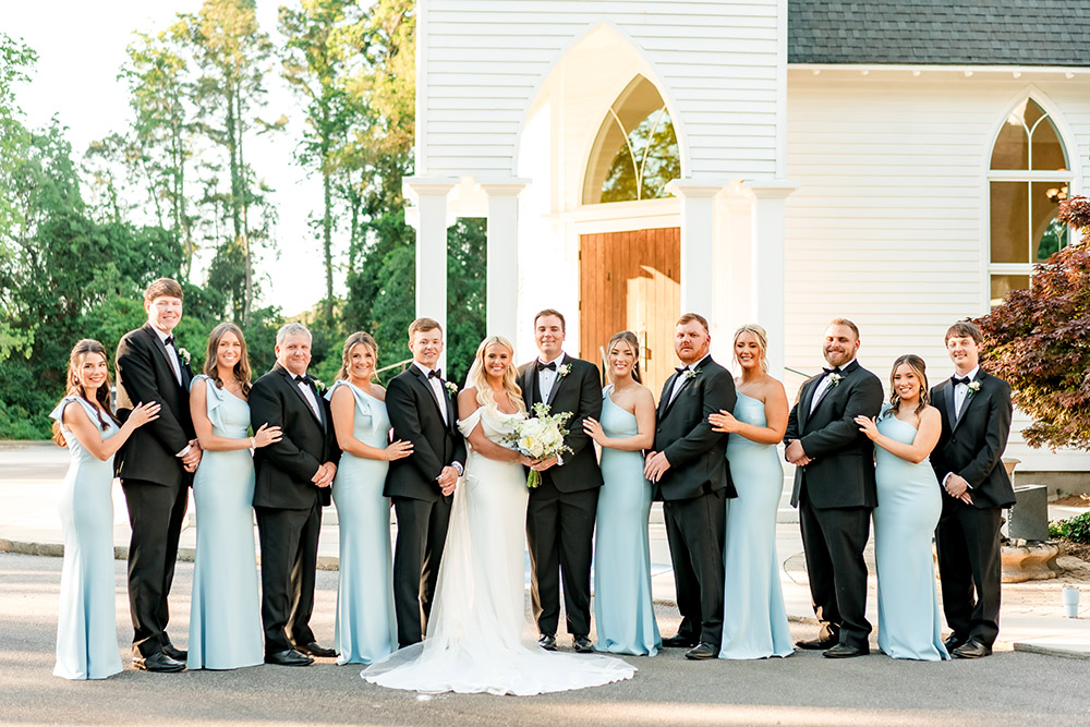 Bride and groom pose with their wedding party and family members outside a white chapel, surrounded by bridesmaids in pale blue dresses and groomsmen in black tuxedos.