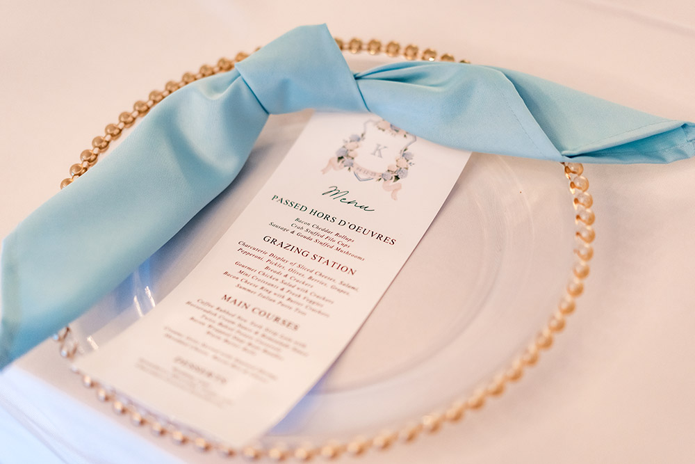 Reception place setting with a pearl-edged charger plate, blue linen napkin, and custom floral menu card.
