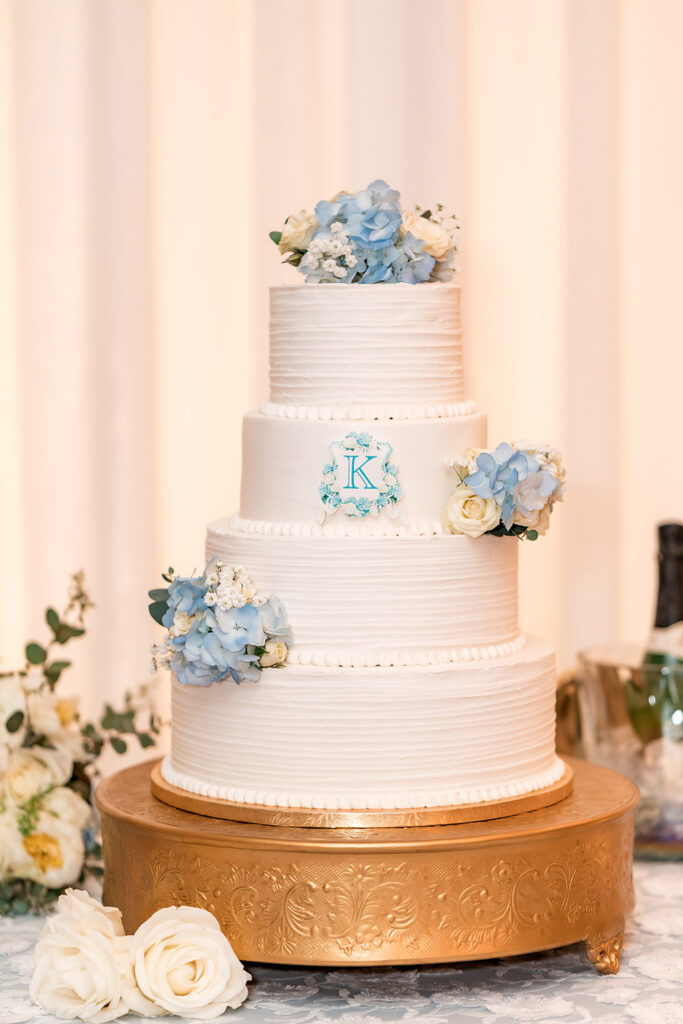 Four-tier white wedding cake decorated with pale blue hydrangeas, white roses, and a custom monogram, displayed on an ornate gold cake stand.