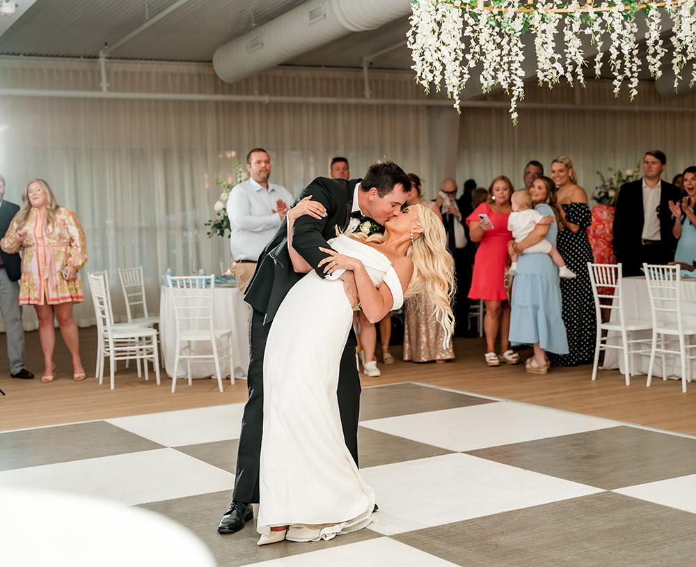 Bride and groom share a dramatic dip kiss during their first dance while family and friends gather around the dance floor.