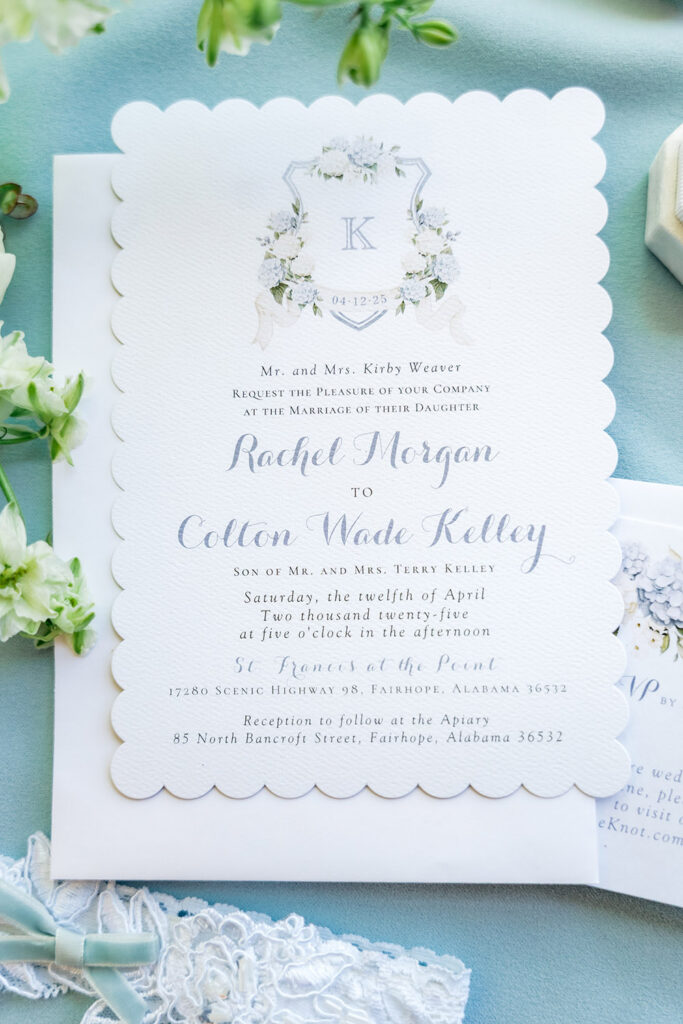 Blue and white floral wedding invitation with scalloped edges styled on pale blue fabric alongside lace, stationery, and fresh flowers.